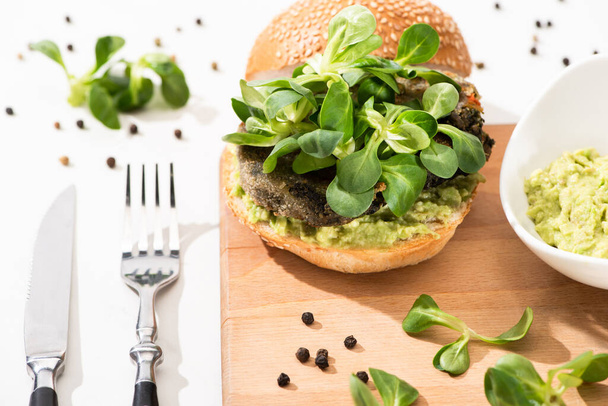 delicious green vegan burger with microgreens and mashed avocado on wooden boar with black pepper near fork and knife on white background - Photo, Image