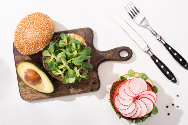 top view of vegan burgers with microgreens, avocado, radish on wooden cutting board on white background with cutlery - Photo, Image