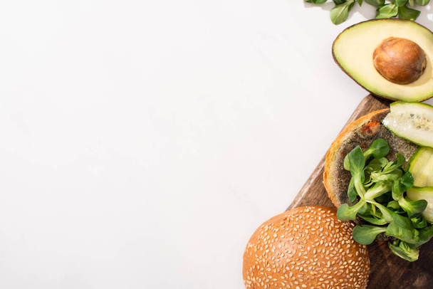 top view of vegan burger with microgreens, cucumber and avocado on wooden cutting board on white background - Photo, Image
