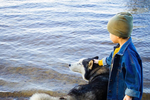 Мальчик гладит большую собаку у реки. Собака с удовольствием закрыла глаза. Настоящие друзья вместе. Вид сбоку. Дневной свет, копировальное пространство
. - Фото, изображение