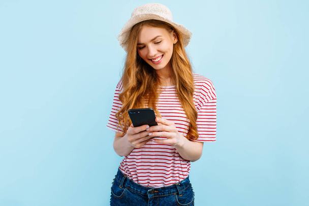 Happy beautiful girl in a summer hat, uses a mobile phone, standing on isolated blue background - Foto, Imagem