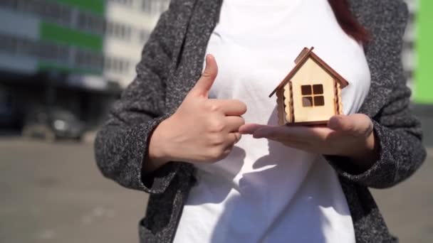 Close-up van de vrouw die kleine houten huis in de hand en andere hand tonen duim omhoog op de achtergrond van gebouwde huis. Concept van de aankoop van een nieuw appartement. - Video