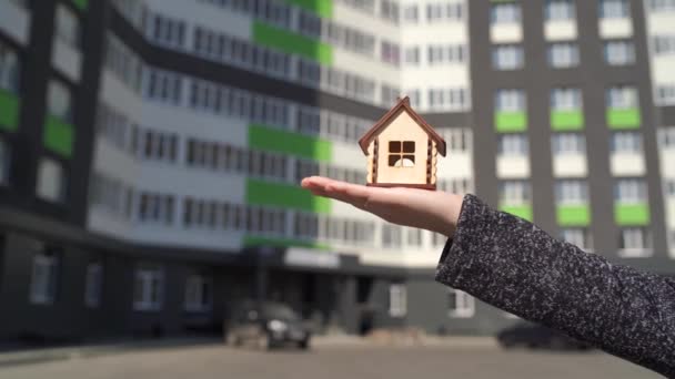 女性の手に小さな木造の家の近くに建てられた家の背景にあります。新しいアパートの購入の概念. - 映像、動画