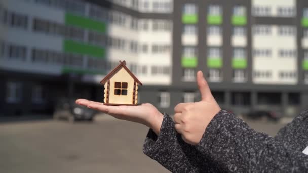 Close up of woman holding small wooden house in hand and other hand showing thumb up on background of built house. Concept of purchasing new apartment. - Footage, Video
