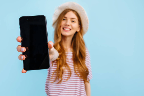 Happy beautiful girl in a summer hat shows a blank screen mobile phone for copy space, on a blue background - Foto, Bild