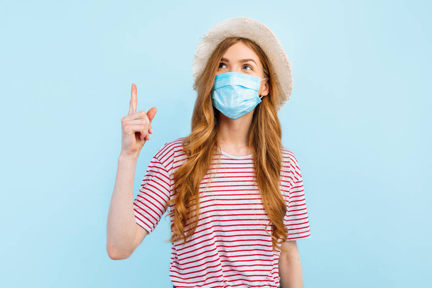 A young woman in a summer hat wears a medical protective mask on her face and points her finger to a copy space on a blue background. The concept of quarantine, coronavirus, summer vacation - Φωτογραφία, εικόνα