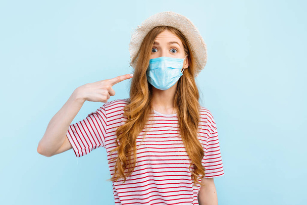 A girl in a summer hat wears a medical protective mask on her face on a blue background. The concept of quarantine, coronavirus, summer vacation - Foto, Bild