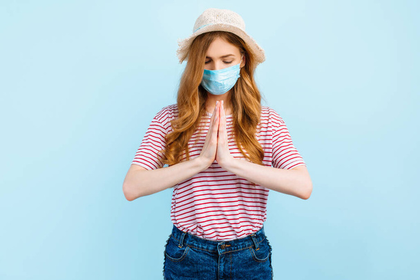 A praying girl in a summer hat wears a medical protective mask on her face, prays, folding her hands together, on a blue background. The concept of quarantine, coronavirus, summer vacation - Photo, Image
