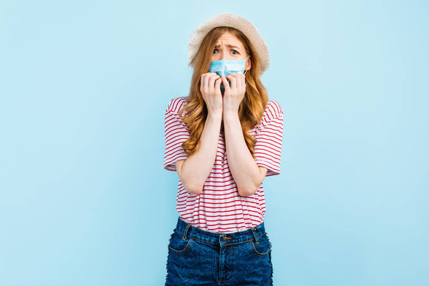 Frightened alarmed girl in a summer hat and in a protective medical mask is afraid and panics, standing on a blue background. Coronavirus concept - Foto, immagini