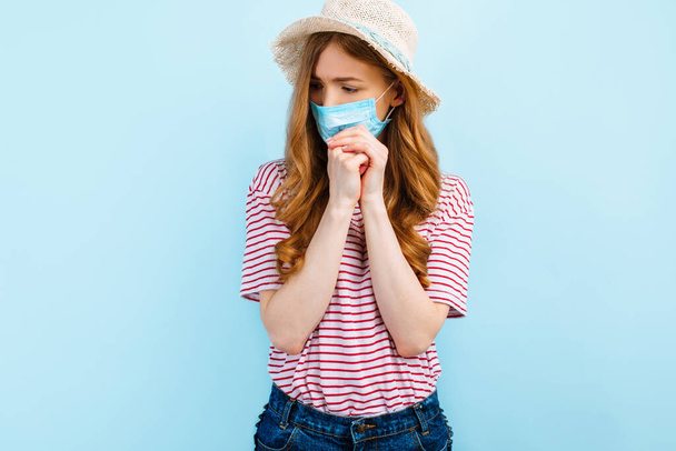 Ein betendes Mädchen mit Sommermütze trägt eine medizinische Schutzmaske auf dem Gesicht, betet mit gefalteten Händen vor blauem Hintergrund. Quarantäne, Coronavirus, Sommerurlaub - Foto, Bild