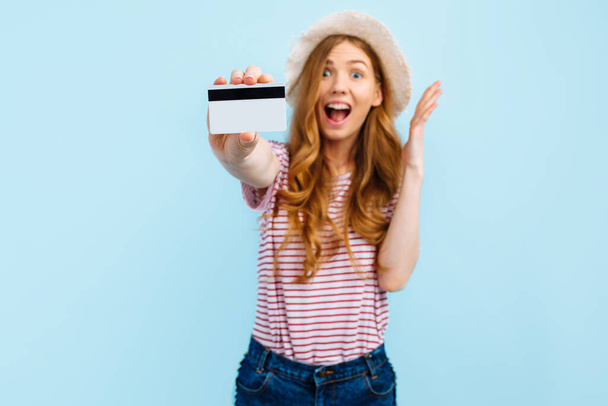 Shocked girl in a summer hat holds and shows a credit card, on a blue isolated background - Foto, afbeelding