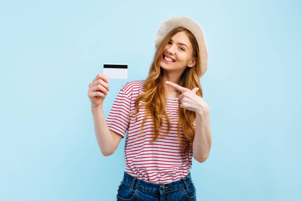 Portrait of a lovely young woman in summer hat showing a credit card and looking into copy space isolated over blue background - Valokuva, kuva