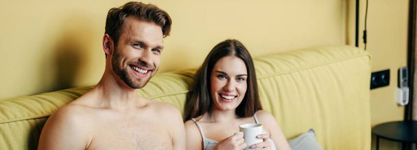 horizontal crop of happy muscular man and cheerful woman looking at camera while holding cups with coffee - Фото, изображение