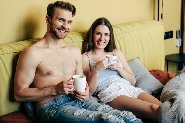 happy muscular man and cheerful woman looking at camera while holding cups with coffee - Foto, immagini