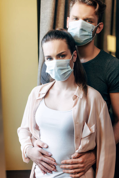 boyfriend hugging girlfriend in medical mask and looking at camera  - Φωτογραφία, εικόνα