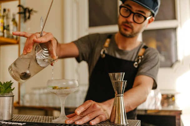 Camarero con delantal, vasos y una tapa que transfiere líquido en un vaso con hielo a una copa de cóctel helada en un elegante bar
 - Foto, imagen