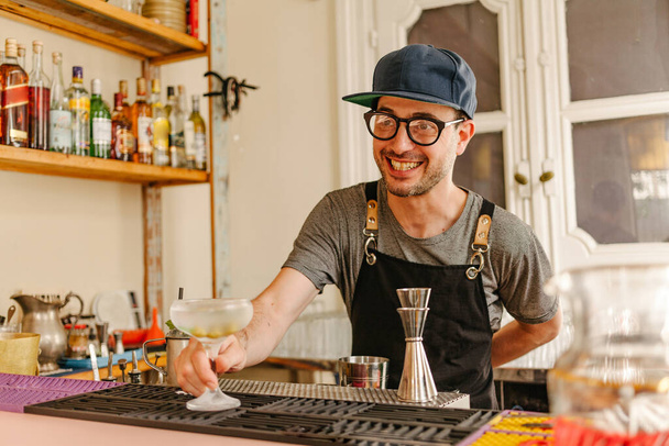 Σερβιτόρος με ποδιά, ποτήρια και καπάκι που δείχνει ένα παγωμένο ποτήρι με ένα κοκτέιλ που παρασκευάζεται στο μπαρ ενός σύγχρονου μπαρ γεμάτο μπουκάλια αλκοόλ - Φωτογραφία, εικόνα
