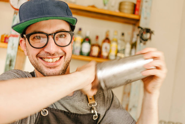 Camarero con delantal, gafas y gorra sonriendo mientras agita un recipiente de cóctel en un bar
 - Foto, Imagen