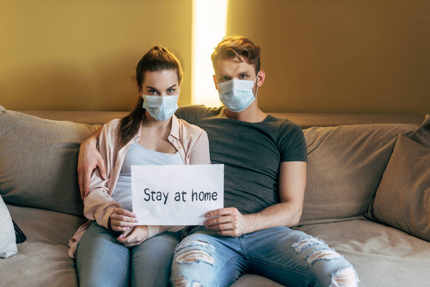 man and woman in medical masks sitting on sofa and holding placard with stay at home lettering  - Foto, Imagen