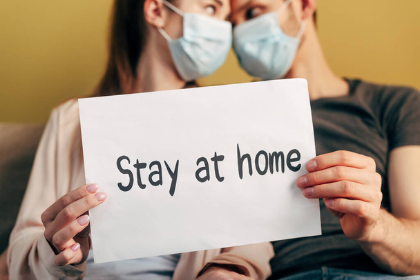 selective focus of man and woman in medical masks looking at each other and holding placard with stay at home lettering  - Zdjęcie, obraz