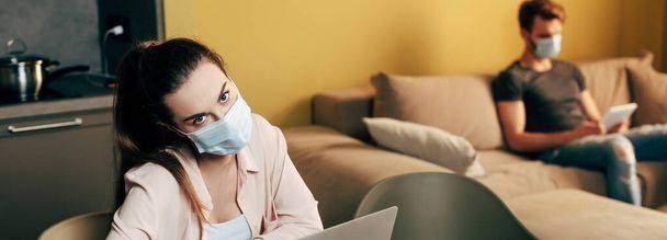 panoramic crop of girl in medical mask looking at camera near man at home  - 写真・画像