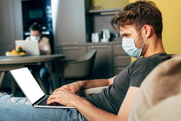 enfoque selectivo de freelancer en máscara médica utilizando el ordenador portátil con pantalla blanca cerca de la novia en casa
 - Foto, Imagen