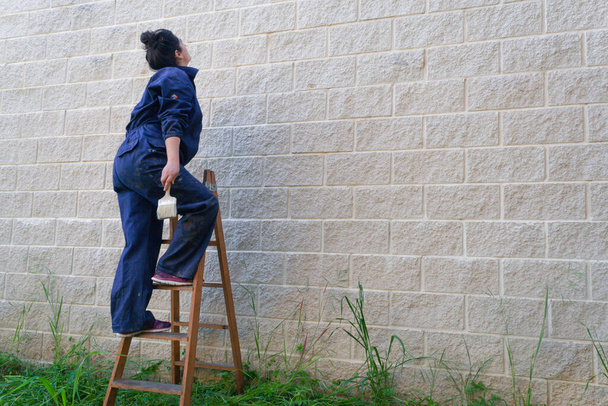 Malarka ubrana w kombinezony robocze wspinająca się po starej drewnianej drabinie na ścianę pędzlem do malowania. - Zdjęcie, obraz