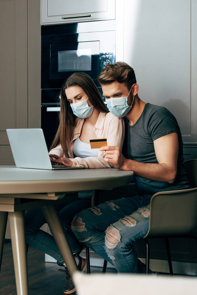 selective focus of man in medical mask holding credit card while sitting near laptop and girl  - Φωτογραφία, εικόνα