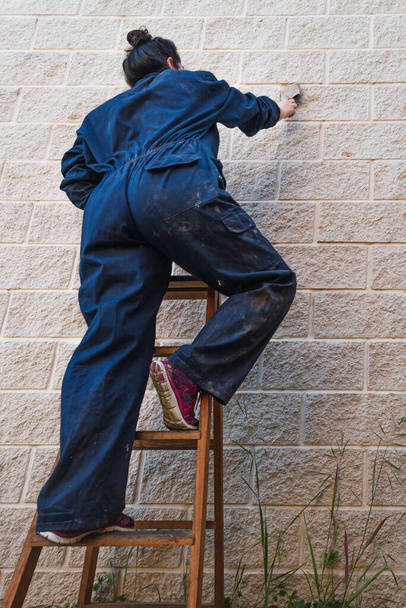 mujer de la pintura vestida con overoles de trabajo escalando una vieja escalera de madera pintando con un pincel. tiro de ángulo bajo. colores suaves
. - Foto, Imagen