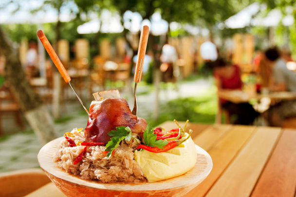 wieprzowina kostki puree ziemniak kapusta chrzan gorący pieprz na drewnianym talerzu w restauracji ogród taras z ludźmi w tle - Zdjęcie, obraz