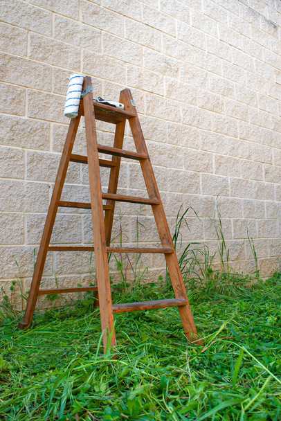 открытая старая деревянная использованная лестница рядом со стеной, на зеленой траве с катком краски
 - Фото, изображение