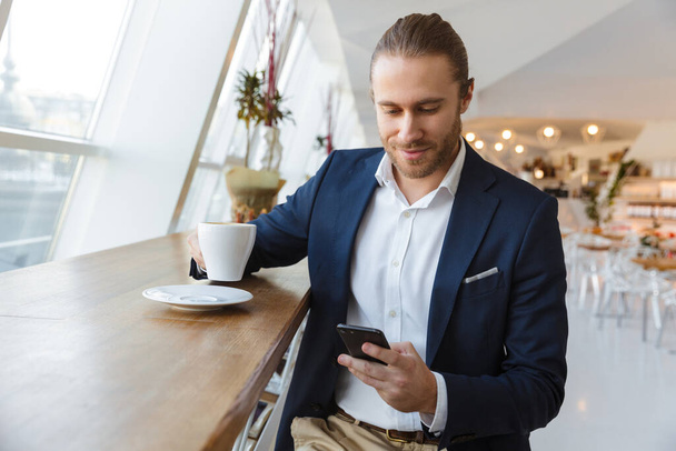 Image of pleased optimistic positive handsome young businessman indoors in office using mobile phone drinking coffee. - Φωτογραφία, εικόνα