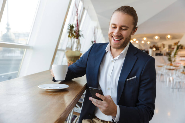 Image of pleased optimistic positive handsome young businessman indoors in office using mobile phone drinking coffee. - Photo, Image
