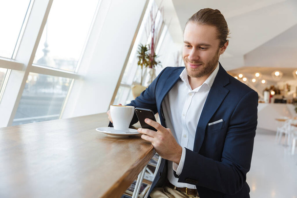 Photo of a cheery smiling handsome young businessman indoors in office using mobile phone. - Foto, Bild