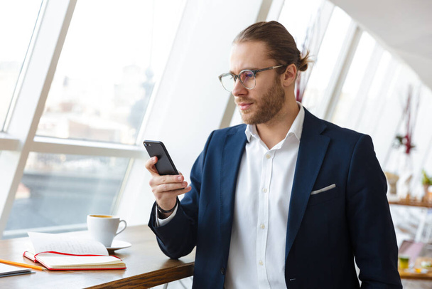 Image of a handsome serious young businessman indoors in office chatting by mobile phone. - Foto, Bild