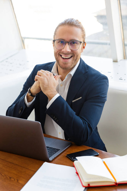 Image of happy positive young businessman indoors in office using laptop computer. - Fotó, kép