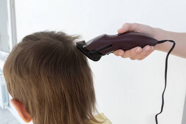 the hand holds an electric hair clipper and cuts the boy 's long hair, hairdresser at home, parent cuts hair while barbershops are closed, stay home concept - Фото, изображение