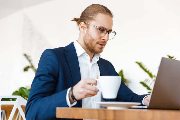 Image of handsome serious young businessman indoors in office using laptop computer drinking coffee. - Foto, afbeelding