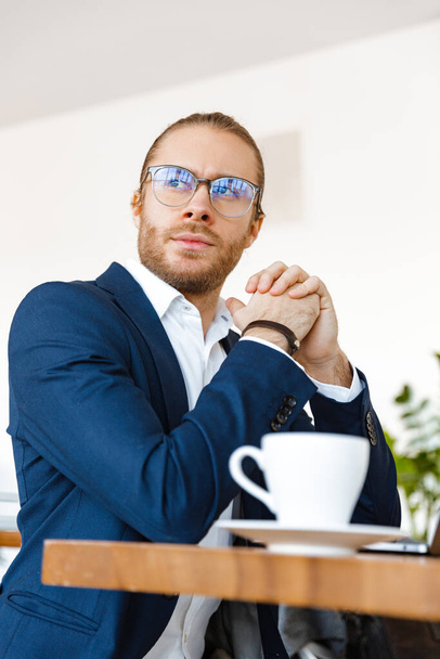 Foto pohledného koncentrovaného mladého podnikatele uvnitř v kanceláři pomocí notebooku pití kávy. - Fotografie, Obrázek