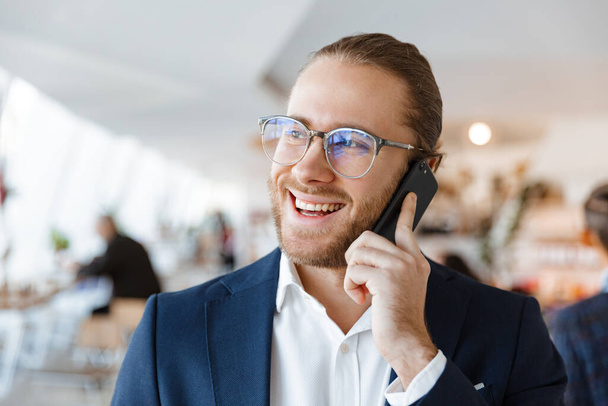 Image of handsome optimistic pleased young businessman indoors in office talking by mobile phone. - Fotoğraf, Görsel