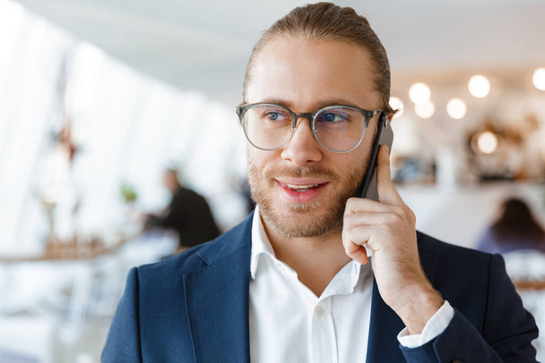 Image of pleased young businessman indoors in office talking by mobile phone. - Фото, изображение