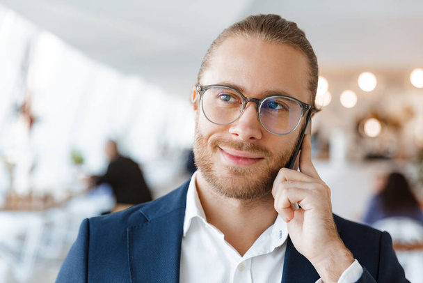 Image of pleased young businessman indoors in office talking by mobile phone. - Photo, Image
