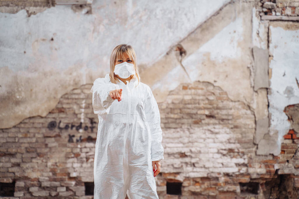 Médica feminina num fato de protecção e máscara médica em pé nas ruínas de uma casa que foi bombardeada. Ela aponta o dedo para a câmara e olha para a frente. Ela está determinada.
 - Foto, Imagem