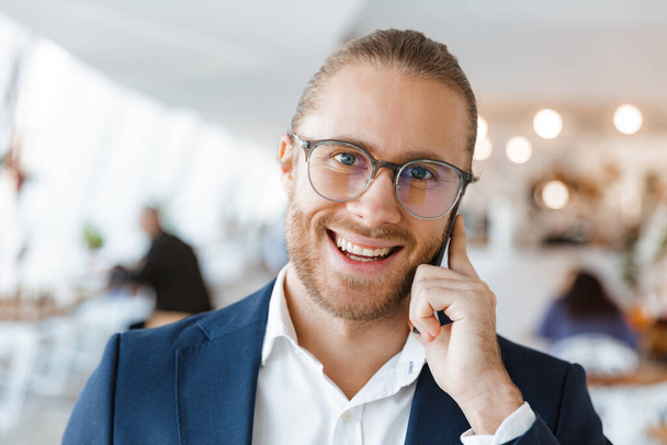 Image of handsome optimistic pleased young businessman indoors in office talking by mobile phone. - Foto, imagen