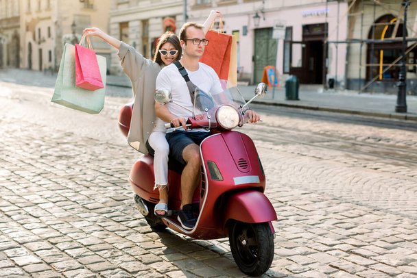 Happy pretty young girl in sunglasses holding arms raised up with colorful shop bags, riding on the motor bike with her handsome boyfriend in the city street with old vintage buildings - Foto, afbeelding