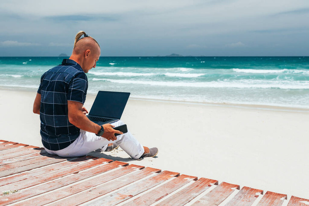 Young guy Holds smartphone in hand. working using a laptop, sitting on the seashore on a sandy beach. Businessman  on a journey. freelancer makes a content work plan - Φωτογραφία, εικόνα