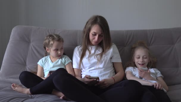 Family is using mobile devices together sitting on sofa during weekend at home. mother and two kids daughter holding smartphones - 映像、動画