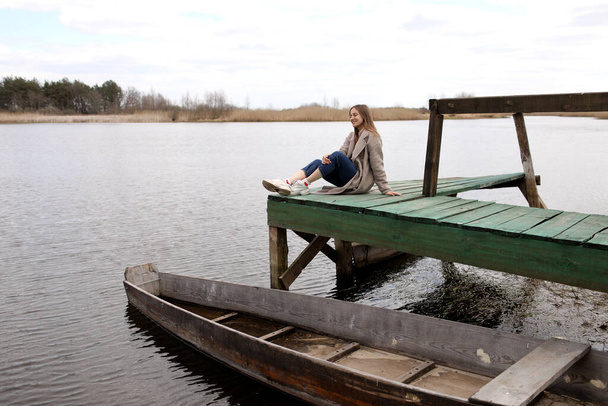 Junge schöne Frau sitzt an einem Frühlingstag auf der Holzbrücke über dem Fluss. altes Boot in Ufernähe. Selektiver Fokus - Foto, Bild