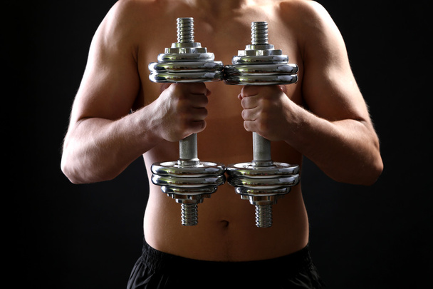 Handsome young muscular sportsman execute exercise with dumbbells on dark background - Фото, зображення