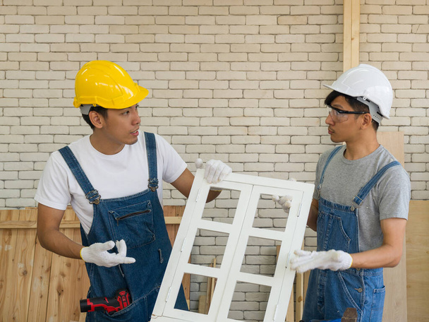 Dos carpinteros asiáticos discutieron sobre las fallas de las ventanas recién ensambladas. Handyman usando un sombrero duro, guantes de tela con un taladro de confianza en la atmósfera de trabajo de la mañana
 - Foto, Imagen